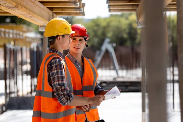 Deux ingénieurs civils vêtus de gilets de travail orange et de casques discutent du processus de construction sur le chantier près des constructions en bois et des cadres en acier — Photo