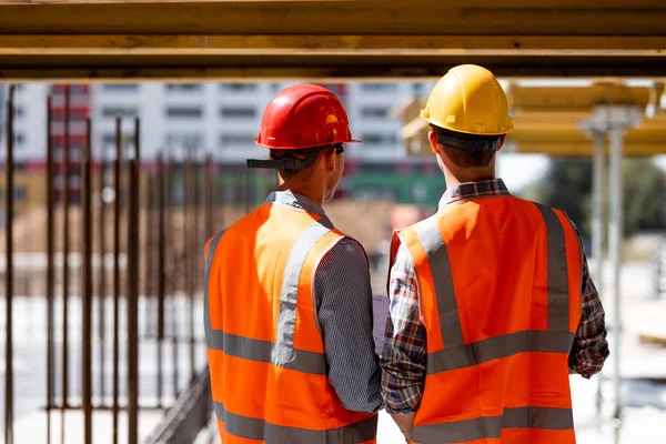 Två civilingenjörer klädd i orange arbete västar och hjälmar diskutera byggprocessen på byggnadsplatsen nära träkonstruktioner och ramar av stål — Stockfoto