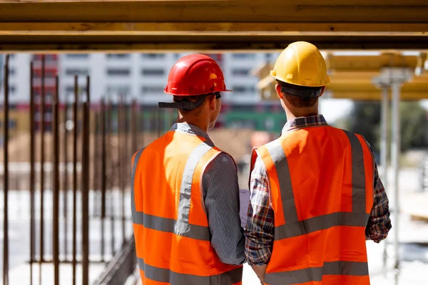 Dois engenheiros civis vestidos com coletes de trabalho laranja e capacetes discutem o processo de construção no local de construção perto das construções de madeira e armações de aço — Fotografia de Stock