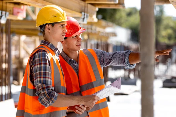 Two civil engineers dressed in orange work vests and helmets discuss the construction process on the building site near the wooden constructions and steel frames — Stock Photo, Image