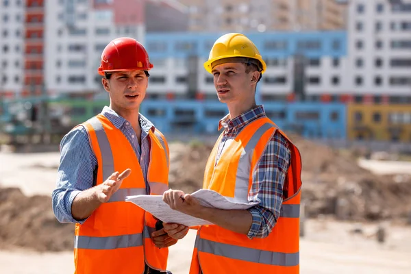 Två män klädda i skjortor, orange arbete västar och hjälmar diskutera dokumentation på byggnadsplatsen mot bakgrund av en flera våningar byggnad — Stockfoto