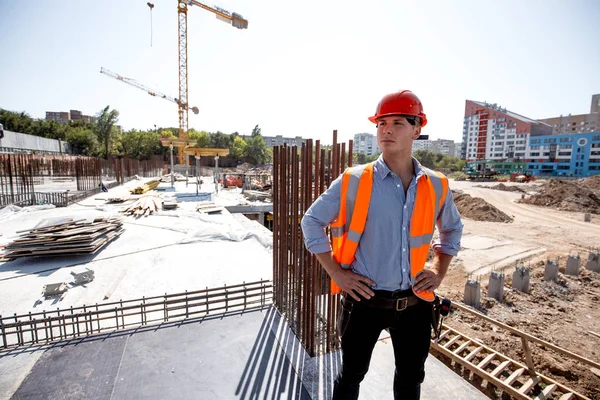 İnşaat alanında bir çok katlı bina arka planı üzerinde gömlek, turuncu iş yelek ve kask giymiş genç adam duruyor