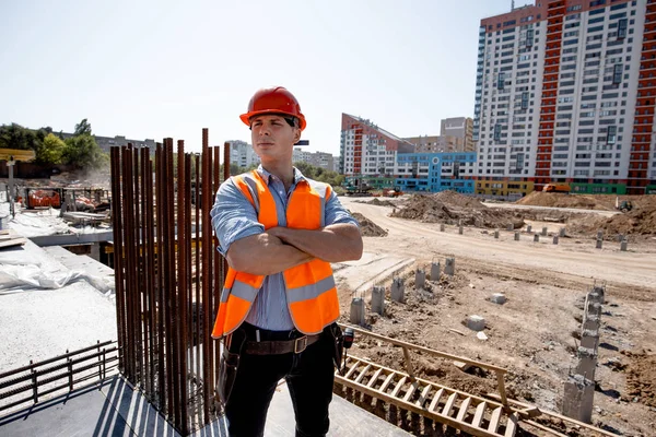 Young man dressed in shirt, orange work vest and helmet stands on the building site against the background of a multi-storey building — Stock Photo, Image