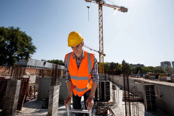 Ung man klädd i skjorta, orange arbetet väst och hjälm fungerar på byggplatsen — Stockfoto