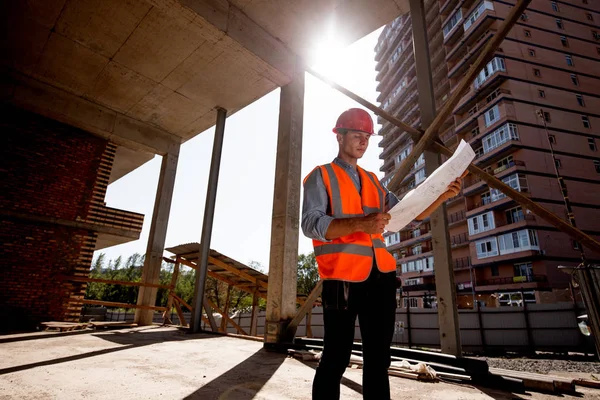 Jeune homme vêtu de chemise, gilet de travail orange et casque explorer la documentation de construction sur le chantier à l'intérieur du bâtiment en construction — Photo
