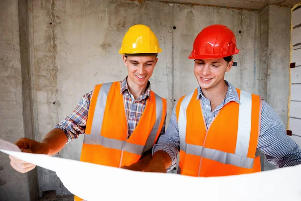 Deux hommes vêtus de gilets de travail orange et de casques explorent la documentation de construction sur un fond de mur en béton — Photo