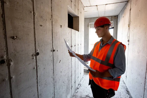 Homme vêtu de gilet de travail orange et casque explore la documentation de construction sur un à l'intérieur du bâtiment en construction — Photo