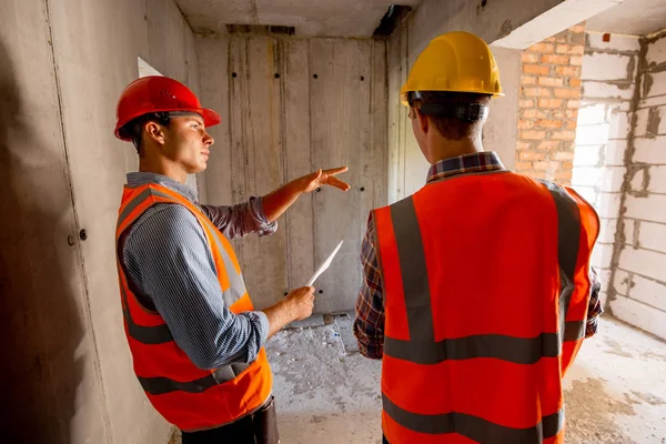 Two civil engineers  dressed in orange work vests and helmets walk inside the building under construction