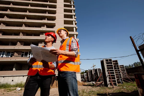 Two structural engineers dressed in shirts, orange work vests and helmets explore construction documentation against the background of a multistorey building