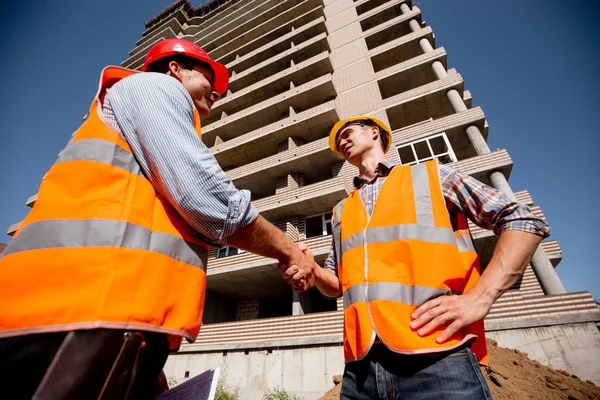 Två män klädda i skjortor, orange arbete västar och hjälmar skaka hand mot bakgrund av en rekreativ byggnad — Stockfoto