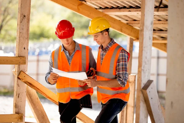 Két férfi öltözött ingek, mellények narancssárga munka és sisakok, és fedezze fel építési dokumentációt az építési telek közel a fa épületszerkezetek — Stock Fotó