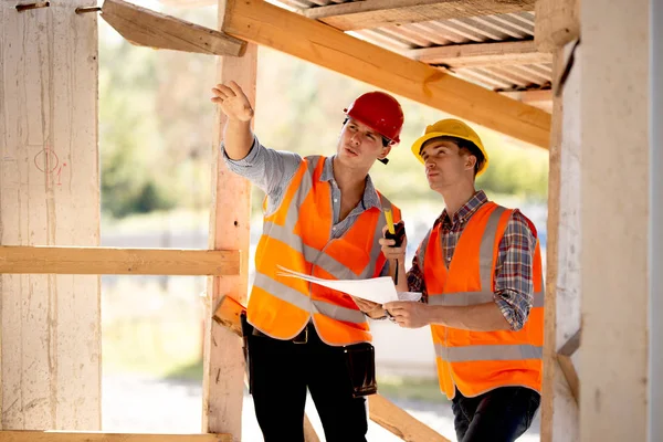Két férfi öltözött ingek, mellények narancssárga munka és sisakok, és fedezze fel építési dokumentációt az építési telek közel a fa épületszerkezetek — Stock Fotó