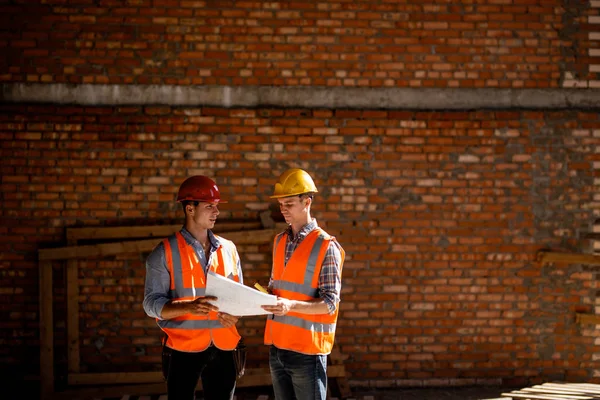 Architecte et directeur de la construction vêtus de gilets de travail orange et casques discutent de la documentation sur un fond de mur de briques — Photo