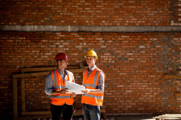 Architect  and construction manager dressed in orange work vests and helmets discuss documentation on a brick wall background