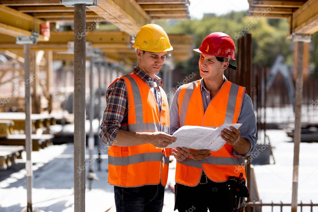 Two civil engineers dressed in orange work vests and helmets explore construction documentation on the building site near the wooden building constructions