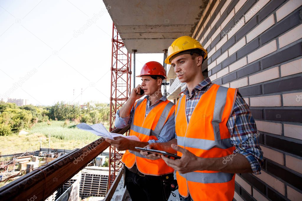 Structural engineer and architect dressed in orange work vests and  helmets discuss the construction process, use a phone and tablet on the balcony of building under construction