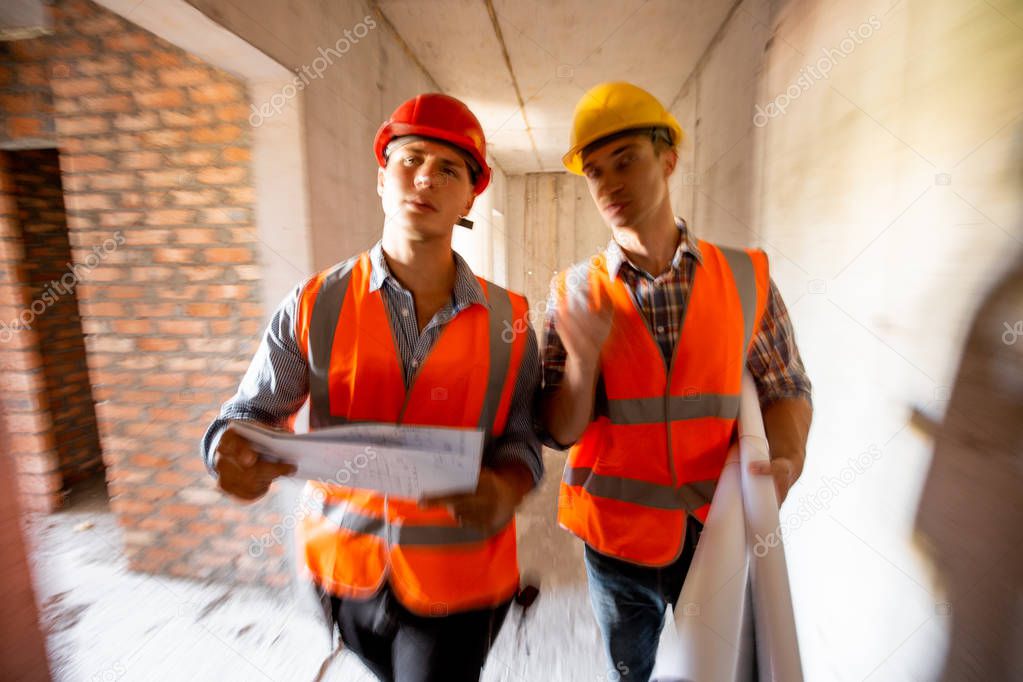 Two civil engineers  dressed in orange work vests and helmets walk inside the building under construction
