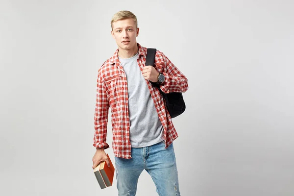 Ung blond kille med svart ryggsäck på hans axel klädd i en vit t-shirt, röd rutig skjorta och jeans håller böcker i sin hand på den vita bakgrunden i studion — Stockfoto