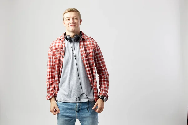 Blond kille med hörlurar på halsen klädd i en vit t-shirt, röd rutig skjorta och jeans står på den vita bakgrunden i studion — Stockfoto