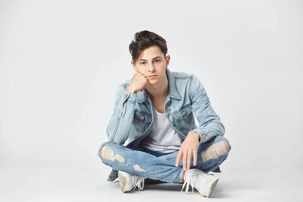 Dark-haired man gekleed in een wit t-shirt, jeans en een denim jasje zit met gekruiste benen op de grond op de witte achtergrond in de studio — Stockfoto