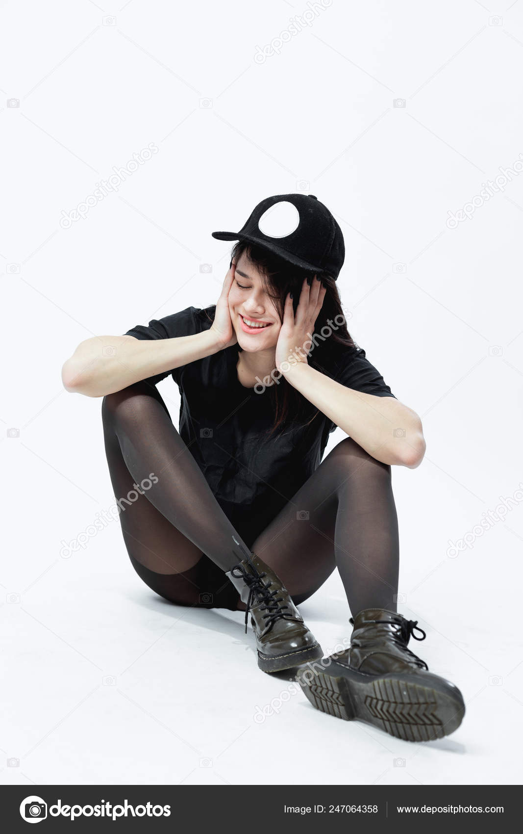 Smiling young dark-haired girl dressed in a black top, shorts, tights and  cap is sitting on the floor on the white background in the studio Stock  Photo
