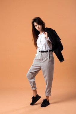 Esmer kız beyaz gömlek, gri pantolon ve siyah spor ayakkabı standları siyah ceketli olan bej renkli arka plan üzerinde stüdyoda omzuna giymiş