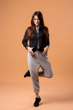 Stüdyoda bej renkli arka plan üzerinde beyaz gömlek, gri pantolon, siyah ceket ve siyah spor ayakkabı giymiş şık siyah saçlı kız poz
