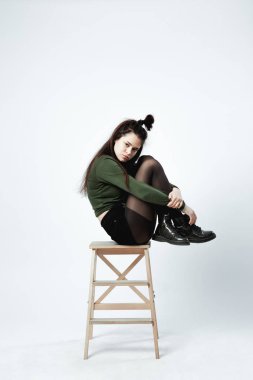 Yeşil gömlek, siyah şort, külotlu çorap ve ayakkabı giymiş komik bir saç modeli ile şık genç esmer kız beyaz arka plan stüdyo ahşap taburede oturan poz