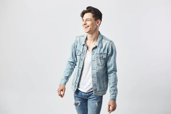 Sorrindo jovem de cabelos escuros cara em uma camiseta branca, jeans e uma jaqueta de ganga fica no fundo branco no estúdio — Fotografia de Stock