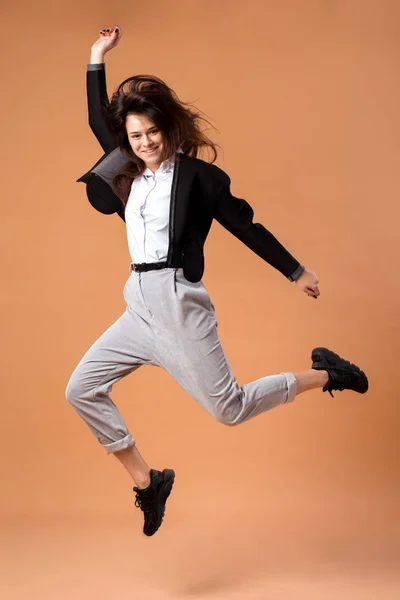 Happy dark-haired girl dressed in white shirt, gray trousers, black jacket  and black sneakers jumps on the beige background in the studio