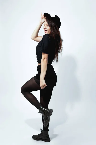 Щаслива стильна темноволоса дівчина, одягнена в чорну футболку, шорти, колготки і кепка, позує на білому тлі в студії — стокове фото