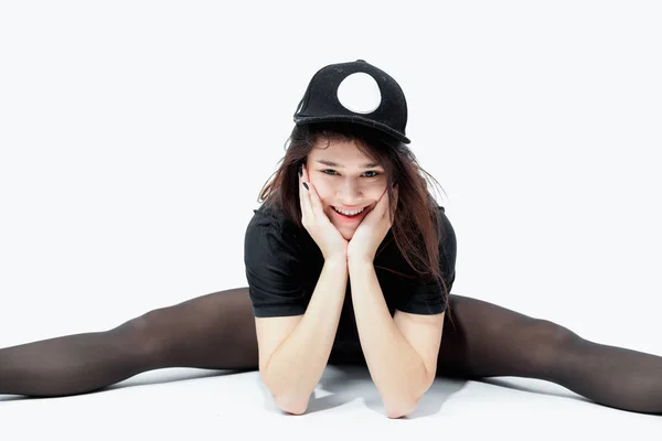 Sorrindo jovem de cabelos escuros menina vestida com um top preto, shorts, collants e boné está sentado no chão no fundo branco no estúdio — Fotografia de Stock