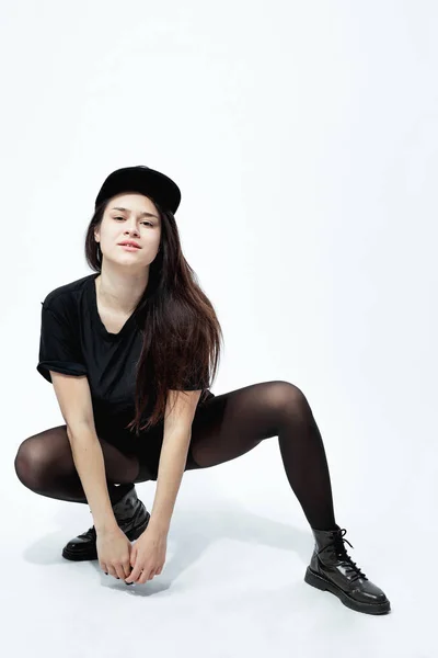 Mladé stylové tmavovlasá dívka oblečená v černý top, šortky, kalhoty a čepici sedí v dřepu na bílém pozadí v ateliéru — Stock fotografie
