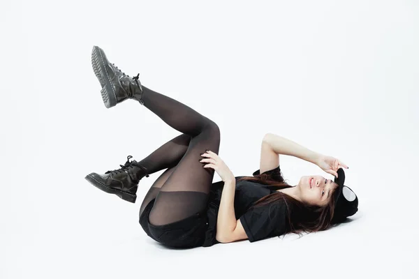 Schlanke junge dunkelhaarige Mädchen in einem schwarzen Top, kurzen Hosen, Strumpfhosen und Mütze liegt auf dem Boden auf dem weißen Hintergrund im Studio — Stockfoto