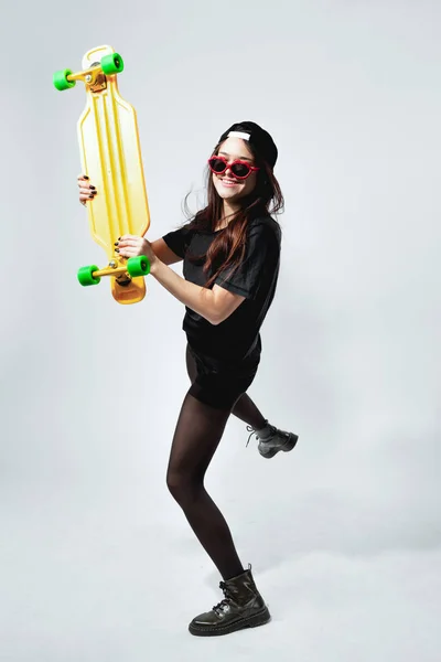 Menina feliz jovem de cabelos escuros em um top preto, shorts, collants, boné e óculos de sol vermelhos com skate amarelo está posando no fundo branco no estúdio — Fotografia de Stock