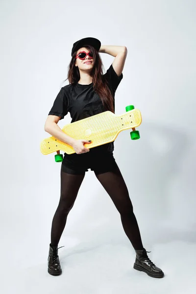 Молода темноволоса дівчина в чорному верху, шорти, колготки, кепка і червоні сонцезахисні окуляри з жовтим скейтбордом позує на білому тлі в студії — стокове фото