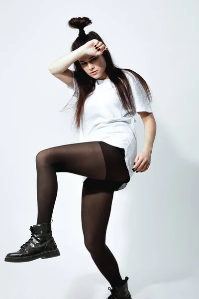 Stylový mladá černovlasá dívka s legrační účes, oblečený v bílé tričko, černé punčocháče a boty je Pózování na bílém pozadí v ateliéru — Stock fotografie