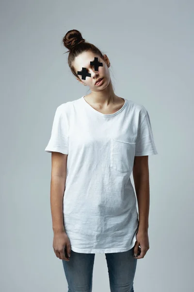 Молода дівчина в білій футболці і джинсах з чорними хрестами клейкої стрічки на очах стоїть на білому тлі в студії, як зомбі — стокове фото