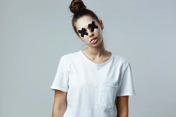 Chica vestida con camiseta blanca con cruces negras de cinta adhesiva en los ojos se hace pasar por un zombie en el fondo blanco en el estudio — Foto de Stock