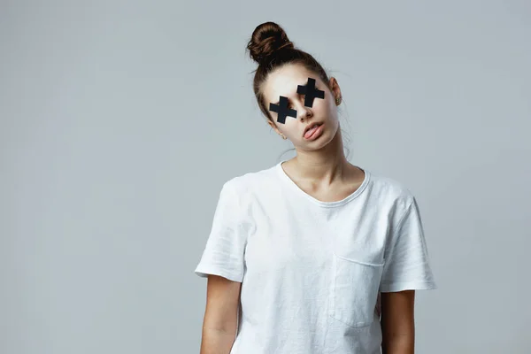 Chica vestida con camiseta blanca con cruces negras de cinta adhesiva en los ojos se hace pasar por un zombie en el fondo blanco en el estudio — Foto de Stock