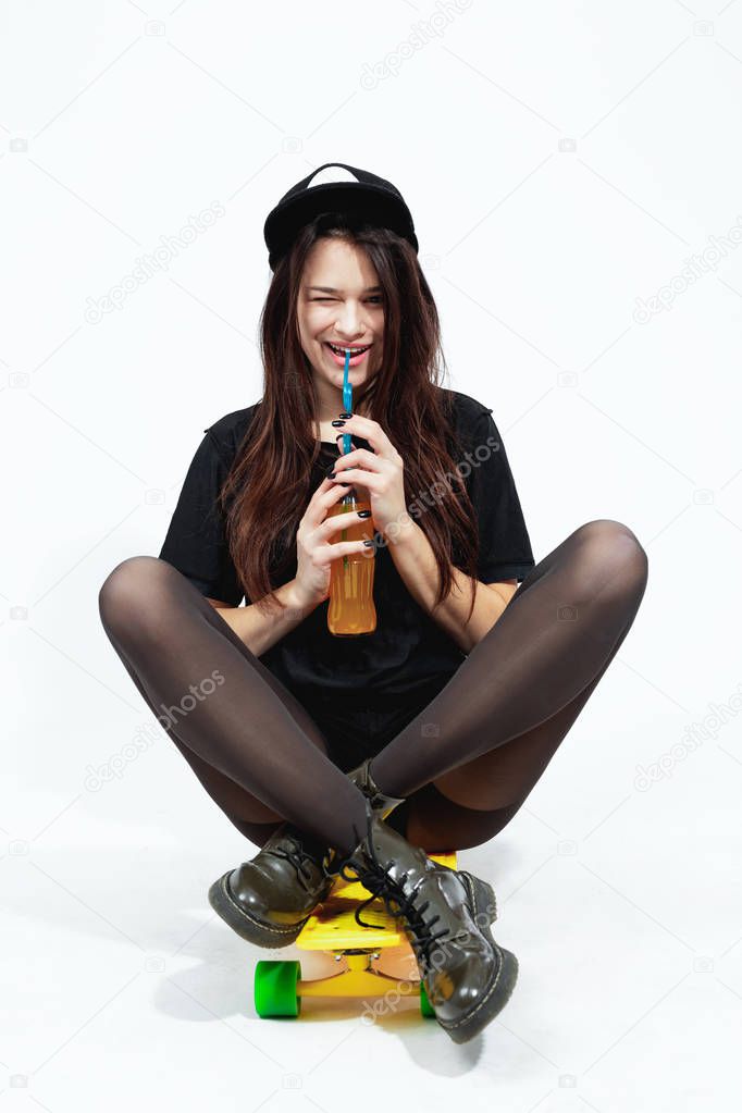 Young dark-haired girl in a black top, shorts, tights, cap and red sunglasses is sitting with a bottle of juice on the floor  in the studio on the white background