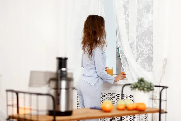 Jonge brunette meisje gekleed in de lichtblauwe pyjama stands met glas vers sap naast het venster in de lichte kamer met het tafeltje met juicer en sinaasappelen — Stockfoto