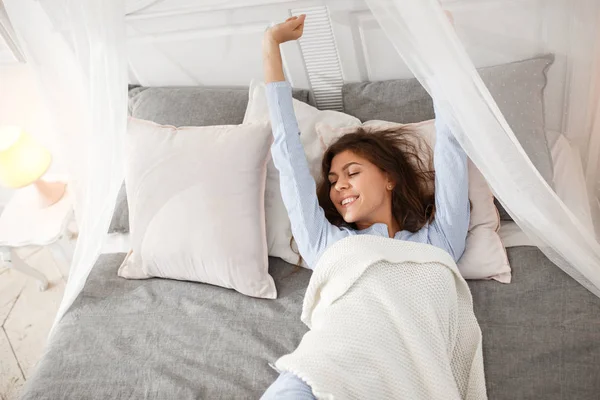 Красива брюнетка дівчина в світло-блакитній піжамі прокидається на ліжку під бежевою ковдрою на сірому листі в спальні — стокове фото