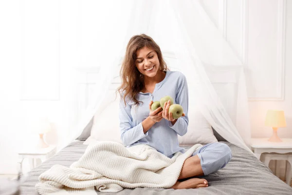 Усміхнена брюнетка в світло-блакитній піжамі сидить на ліжку з зеленими яблуками в руках на сірому листі з бежевою ковдрою — стокове фото