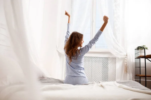 Brunette fille dans le pyjama bleu clair étend ses bras vers le haut assis sur le lit à baldaquin à côté de la fenêtre dans la chambre confortable — Photo