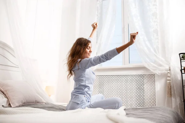 Брюнетка в светло-голубой пижаме вытягивает руки, сидя на кровати с балдахином рядом с окном в уютной спальне. — стоковое фото
