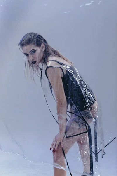 Stylish young girl with wet hair and tattoo on her leg dressed in black swimming suit and transparent rain coat is posing on the white background in the studio