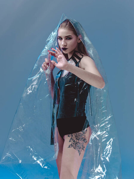 Stylish model with dark lipstick dressed in black swimming suit and transparent rain coat is posing in the transparent membrane in the studio with lighting simulating evening street light