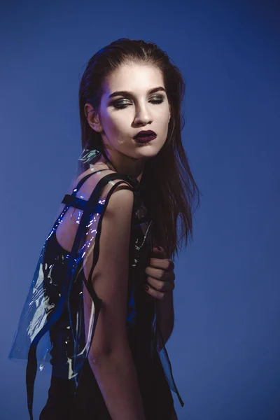 Fashion model with dark lipstick and wet hair dressed in black swimming suit and transparent rain coat is posing in the studio with lighting simulating cold evening street light