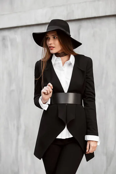 Fashionable slim long-haired girl dressed in a black stylish jacket on a white shirt, black pants and a hat with a wide brim poses in the street — Stock Photo, Image